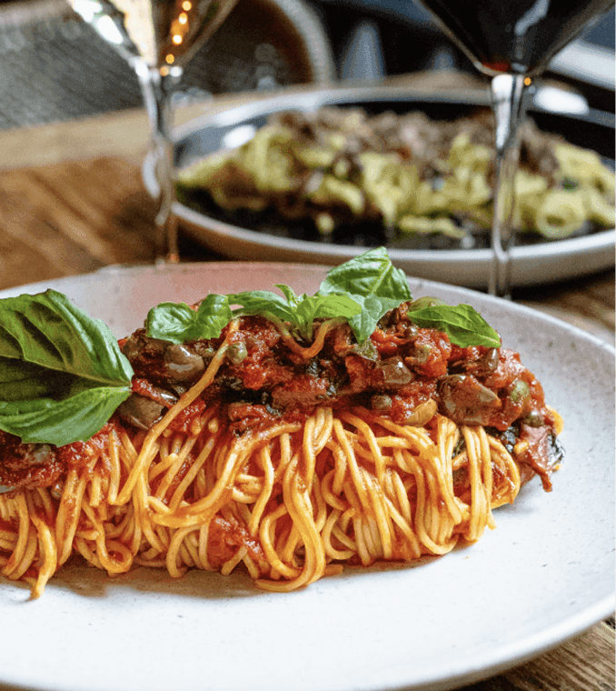A plate of spaghetti topped with tomato sauce, olives, and basil sits on a table with glasses of wine and another pasta dish in the background.