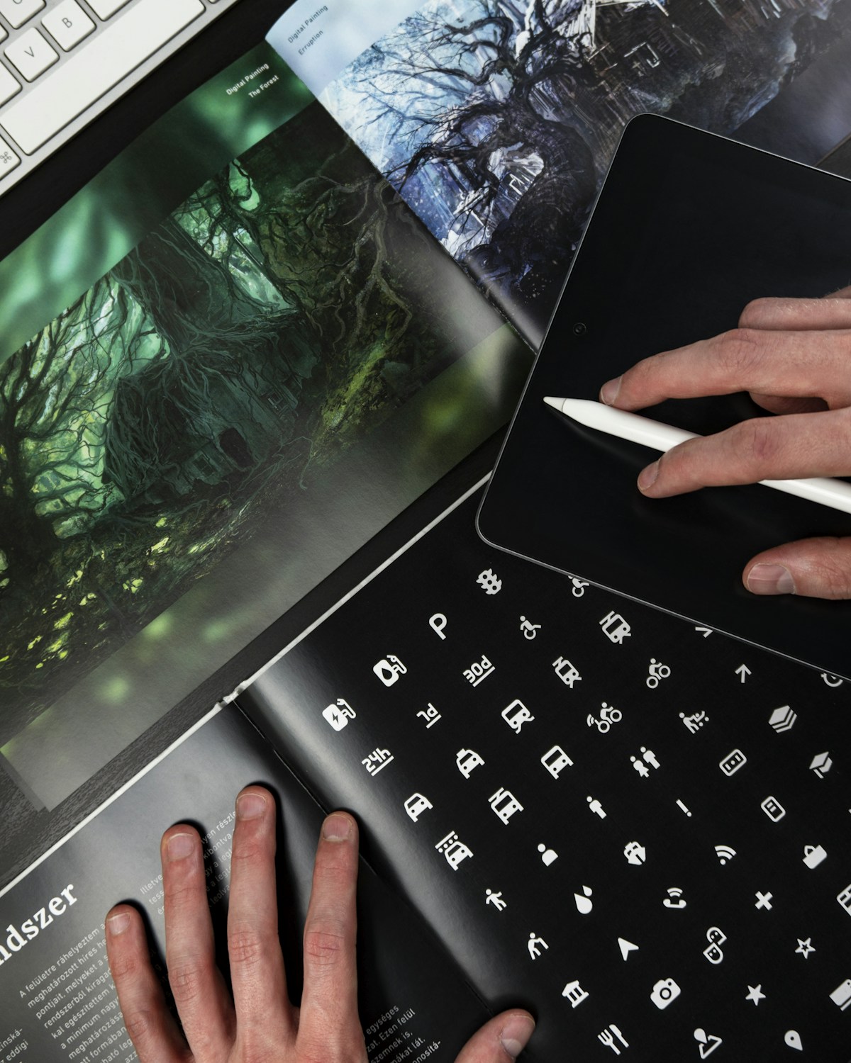 Designer using stylus on tablet next to open magazines with forest art and icons.