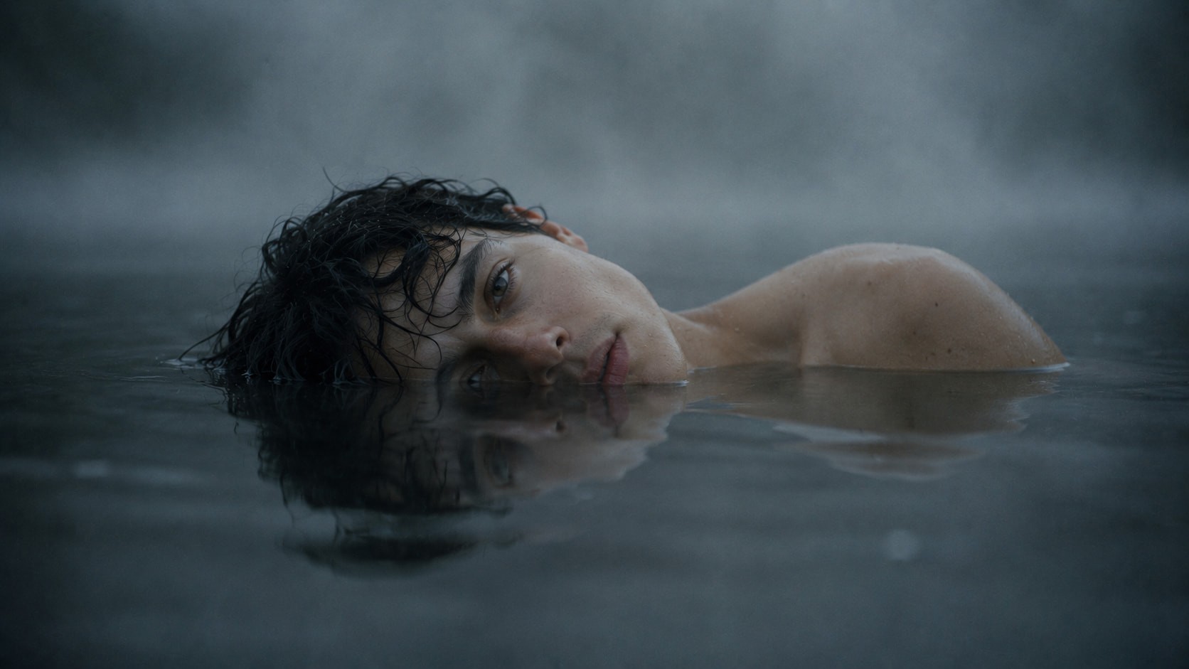 A solitary figure floats at the boundary between submersion and survival, their reflection dissolving into the still water beneath them. The muted tones and direct gaze create a haunting sense of vulnerability, as though they are suspended between surrender and awakening.