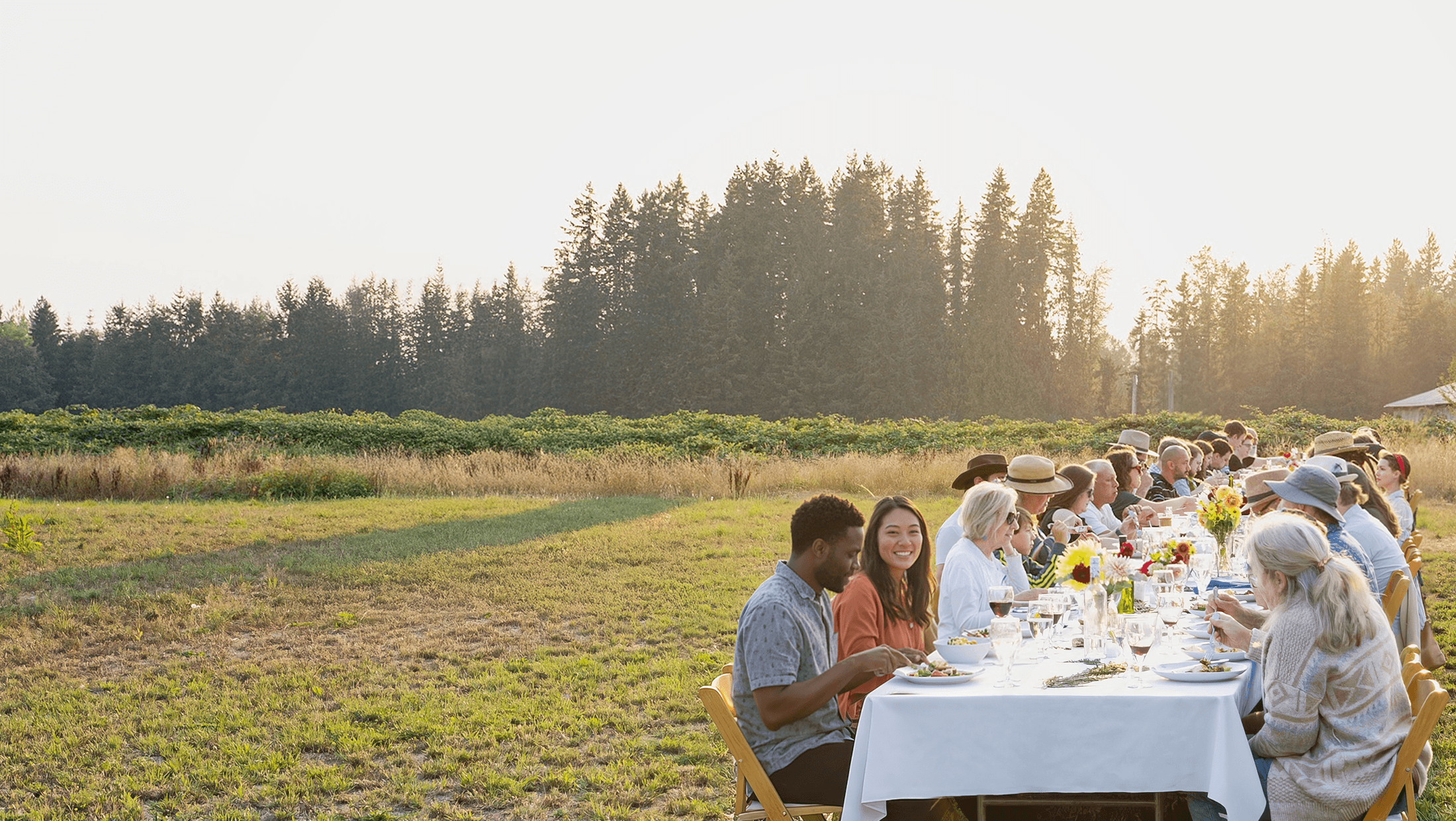 Residents and guests sharing a long outdoor meal together on farmland at Rooted Northwest.