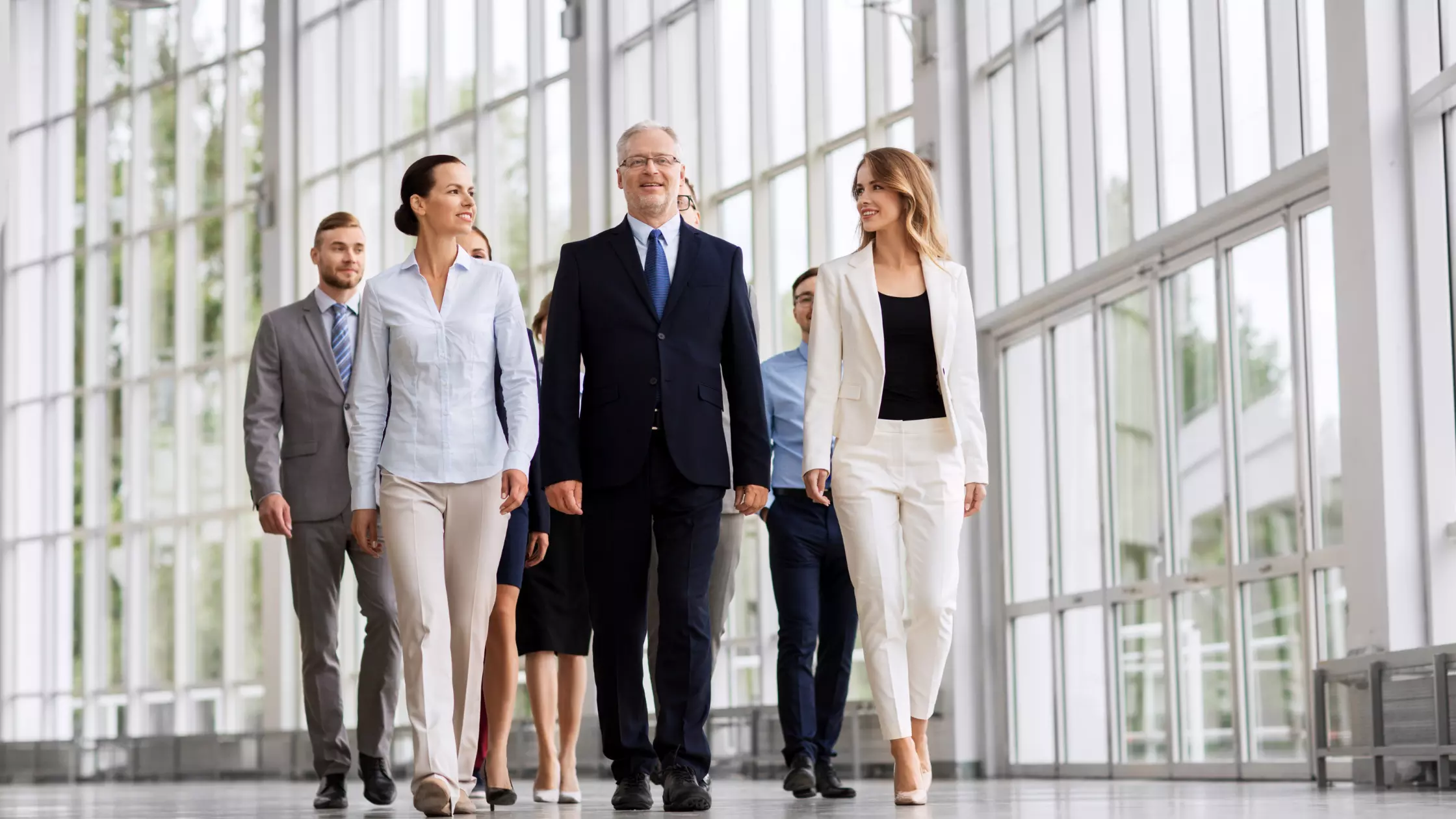 A man stands while two people sit at a table in an office setting, smiling and engaged in conversation. The mood is friendly and collaborative.