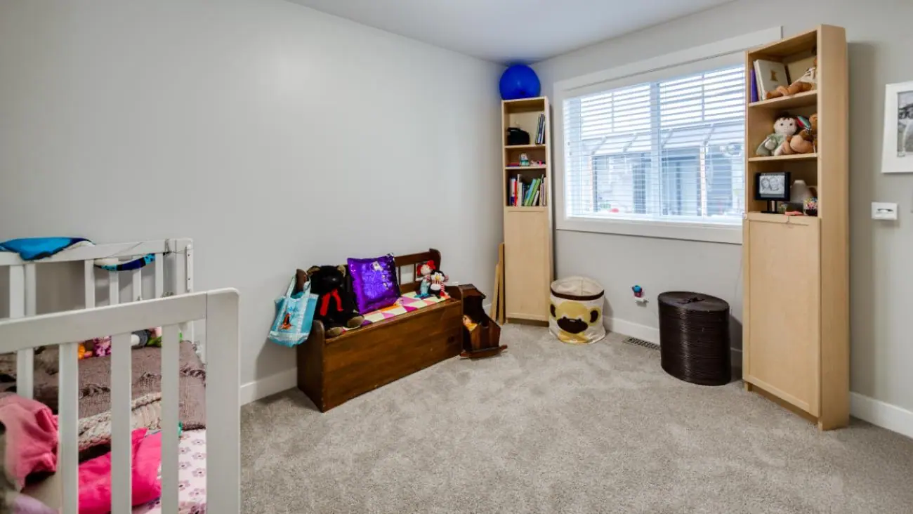 Versatile kids bedroom in multi-generational Burnaby home featuring built-in blonde wood storage shelving system, toy storage solutions, neutral carpet, window seat area, and organized play space design