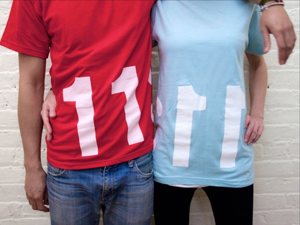 Two people are standing side by side against a white brick wall. The person on the left wears a red t-shirt with the number "11" in white, paired with blue jeans. The person on the right wears a light blue t-shirt, also with the number "11" in white, and black pants. Both individuals have their hands near their hips.