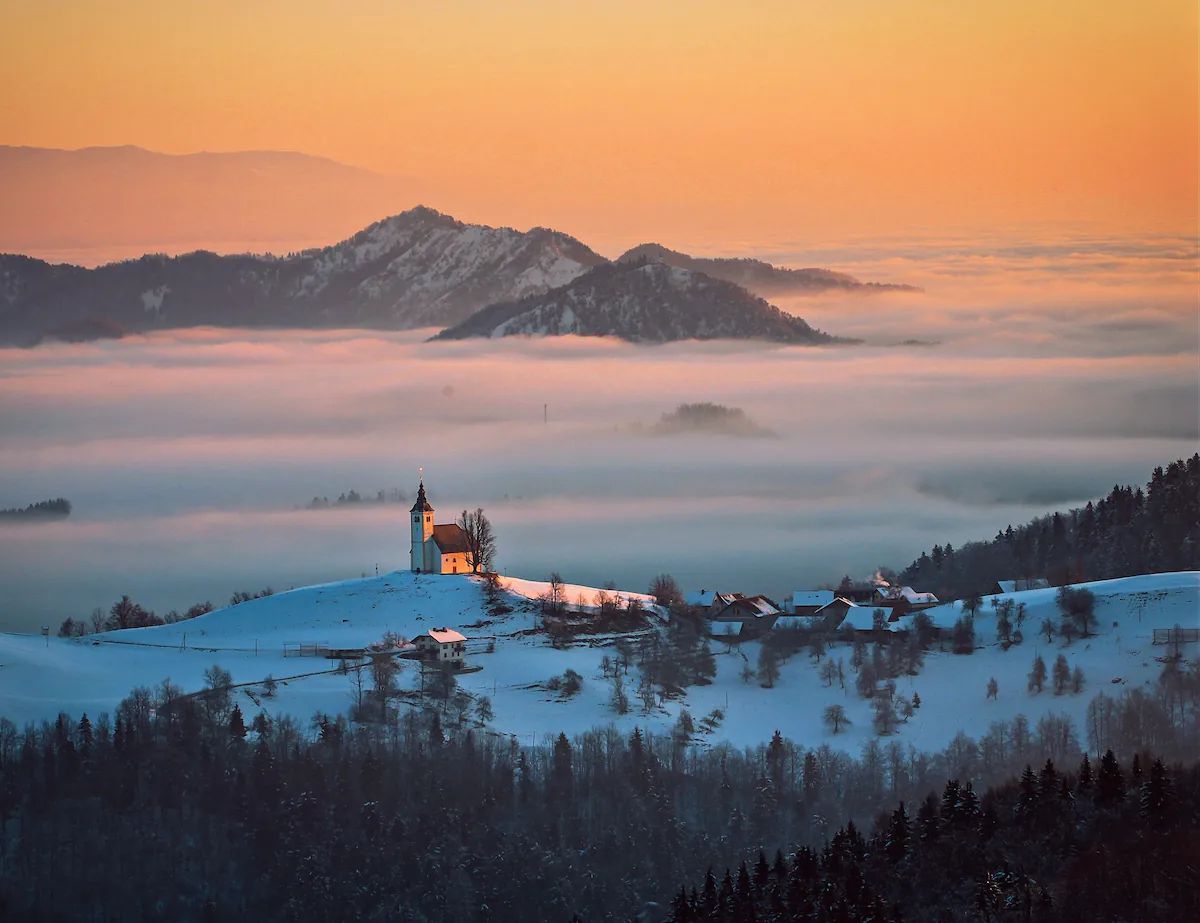 The iconic hilltop church of Sveti Andrej in Slovenia, appearing as an island above a dense layer of low-lying fog during a pastel winter sunrise.
