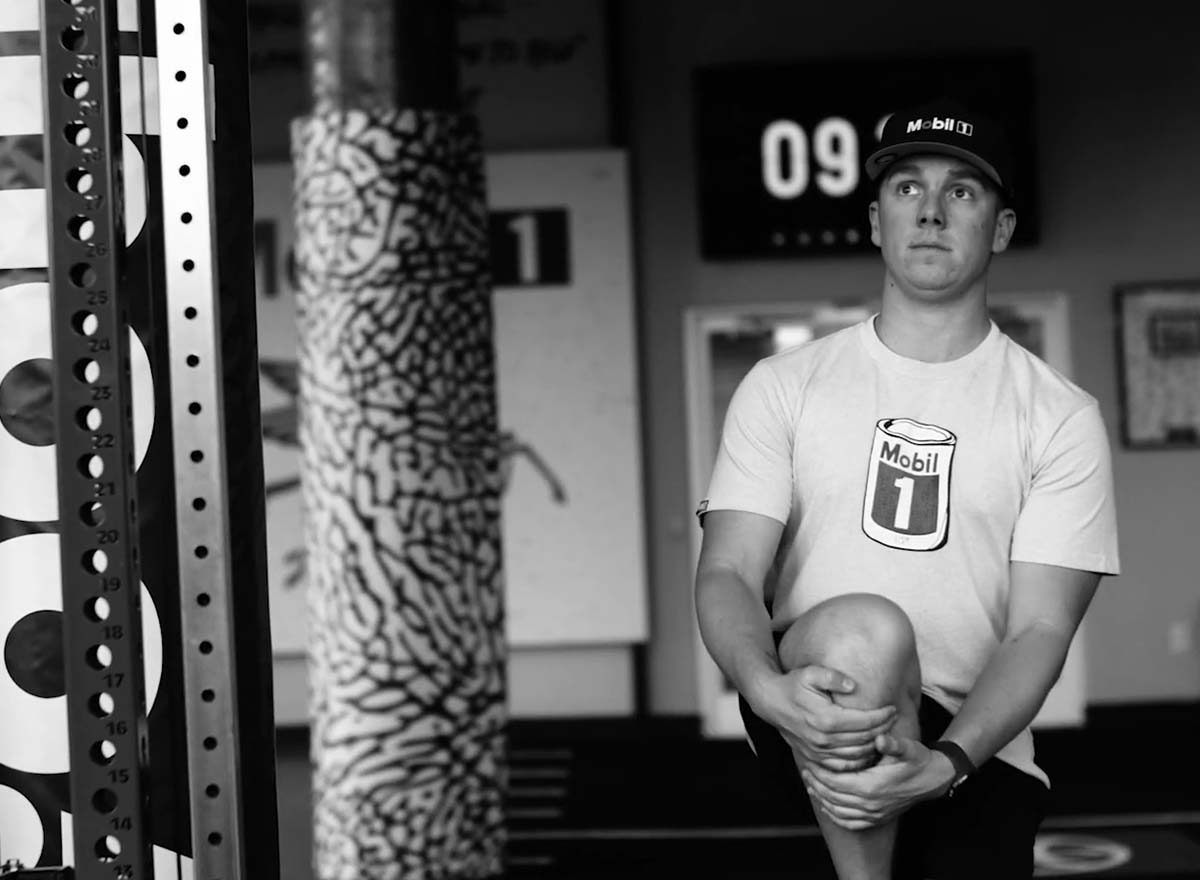 Black-and-white photo of John Hunter Nemechek in a Mobil 1 shirt and hat stretching indoors before a workout.