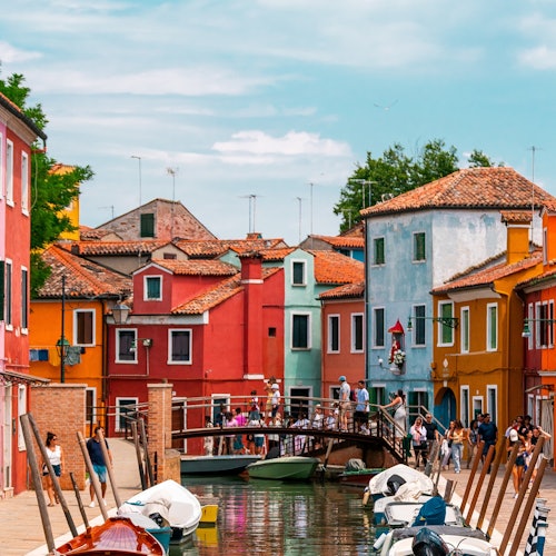 Colorful buildings line a canal with boats docked along the sides. A small bridge with people on it crosses the canal.