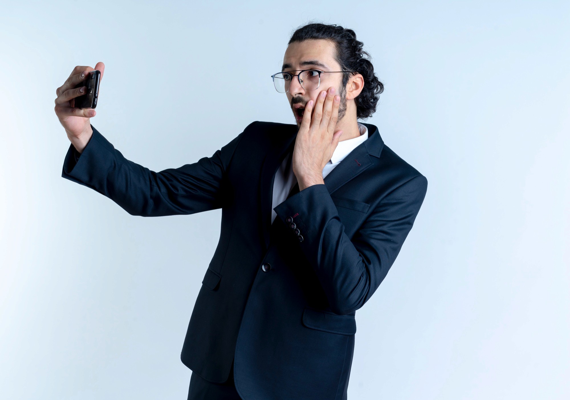 Man in suit taking a surprised selfie, holding phone and touching his face.