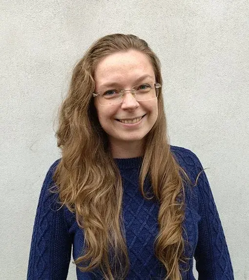 A person with long, wavy brown hair and glasses is smiling, wearing a textured navy blue sweater, standing against a plain light gray background.
