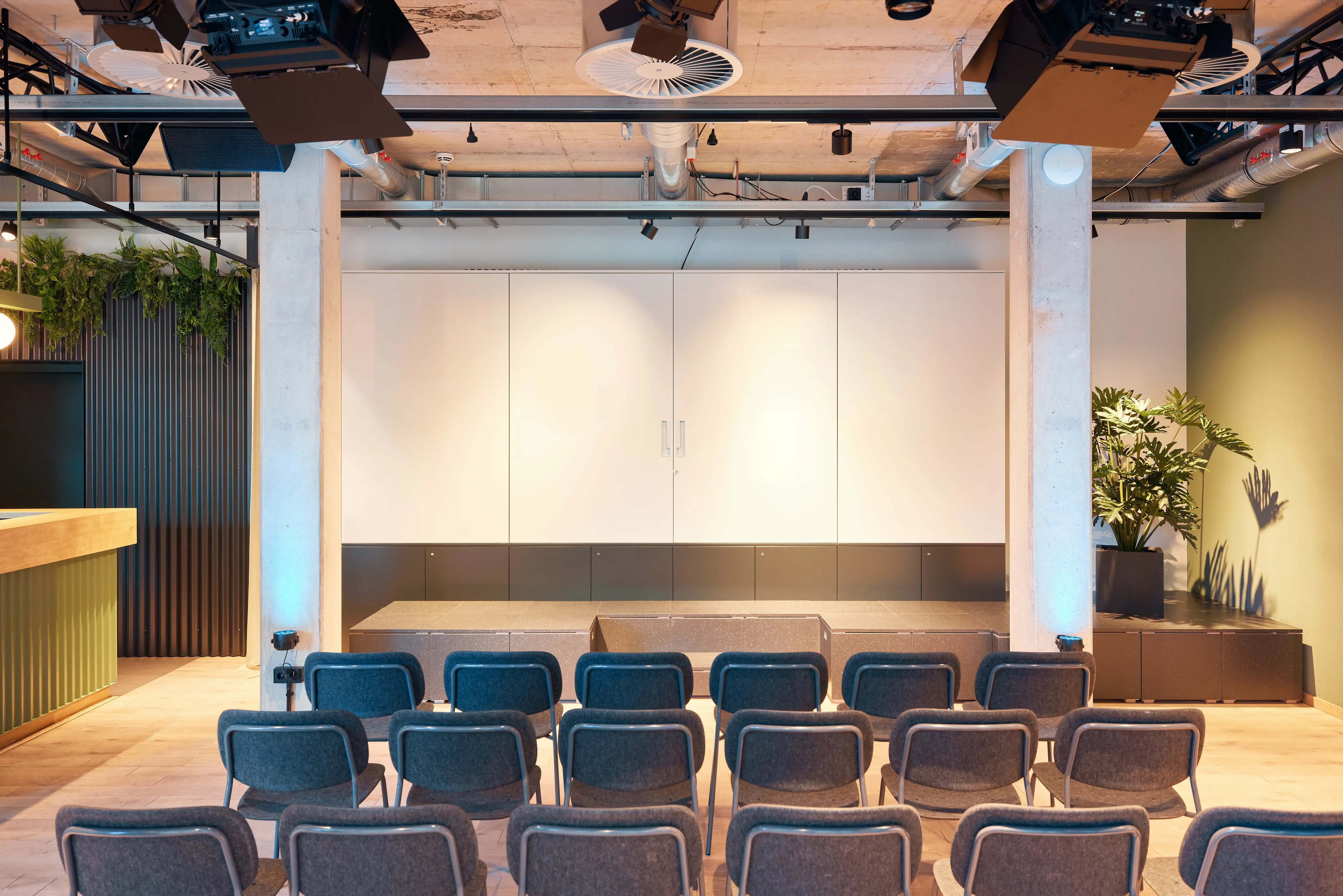 Stage hall at 381 EVENT SPACE with rows of grey upholstered chairs facing a low platform with white wall panels, stage rigging with spotlights and plants on both sides.