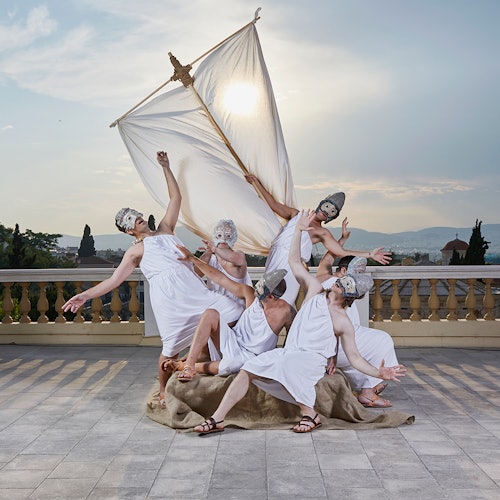 六位身穿托加长袍、戴着面具的人在一面临时搭起的船帆前摆出极具戏剧感的姿势,背景是户外景致与城市天际线。