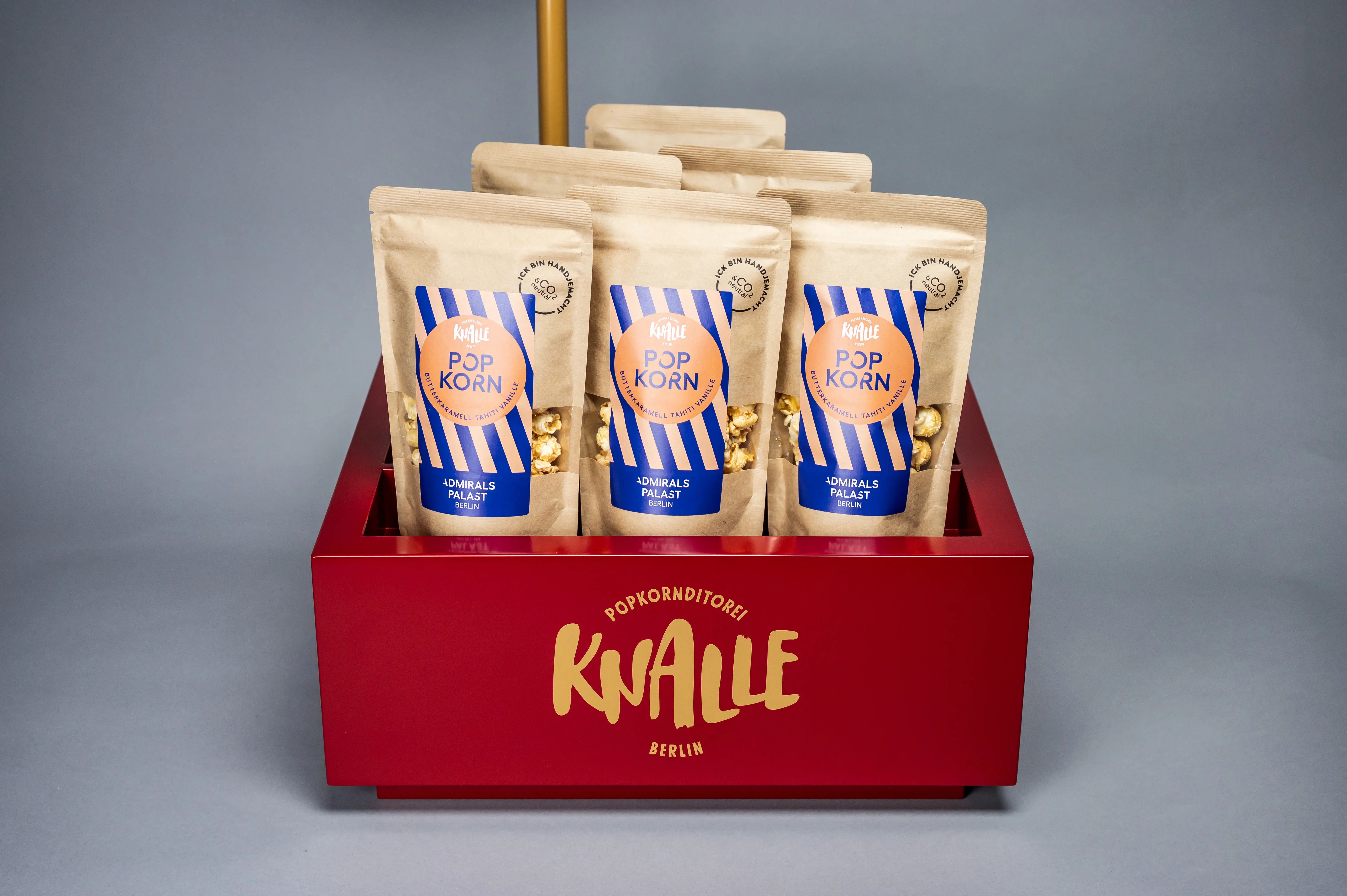 Red KNALLE product tray with golden logo holding striped Popkorn bags of the Admiralspalast edition.