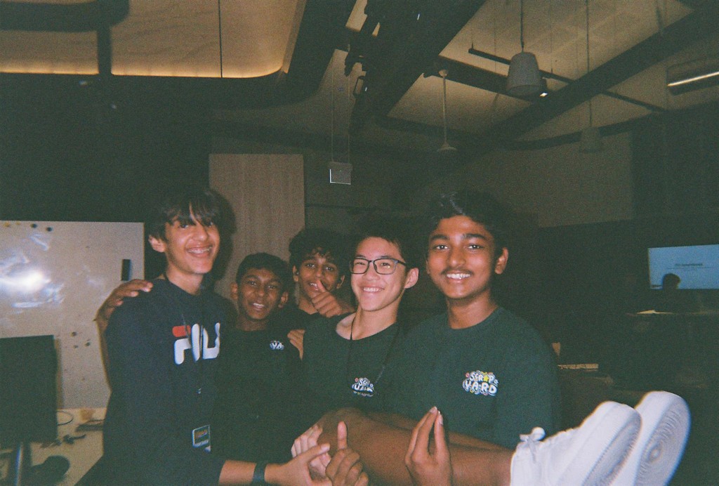 A person wearing headphones sits at a desk with a large curved monitor displaying graphic design software, surrounded by a laptop, smartphone, and potted plant, creating a modern workspace setup.