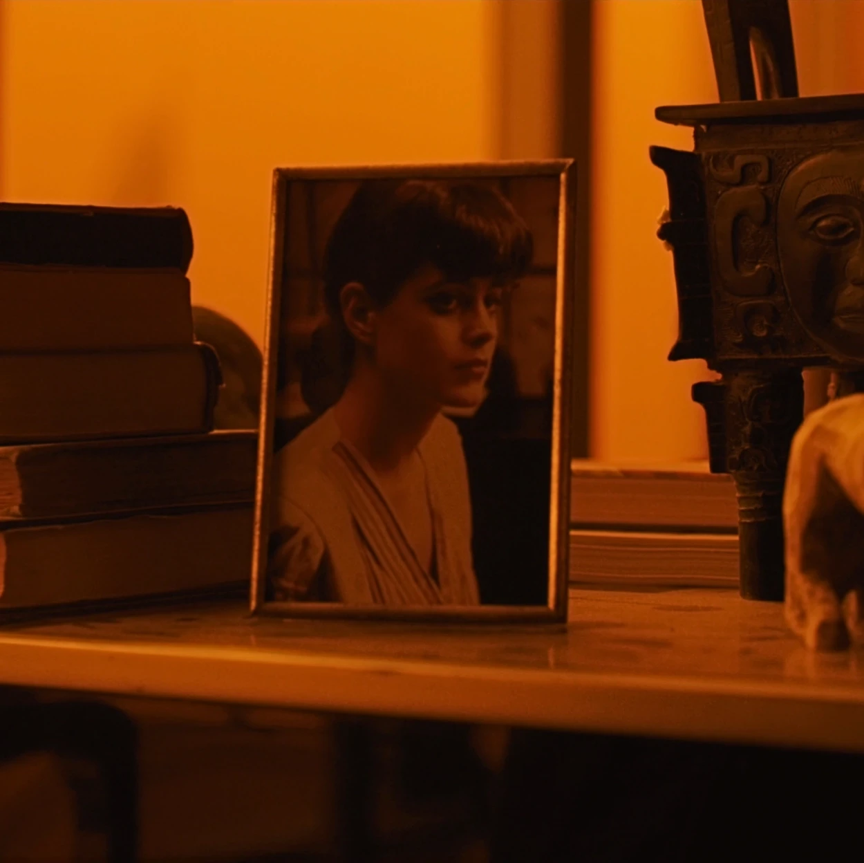 A framed photograph of a woman propped on a wooden surface beside a stack of books and a decorative carved object, bathed in deep orange light