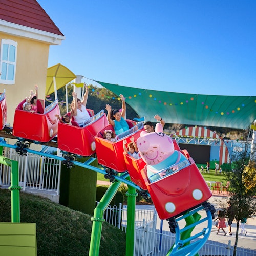 Children and adults ride a roller coaster with red cars shaped like Peppa Pig, against a backdrop of a house and colorful canopy.