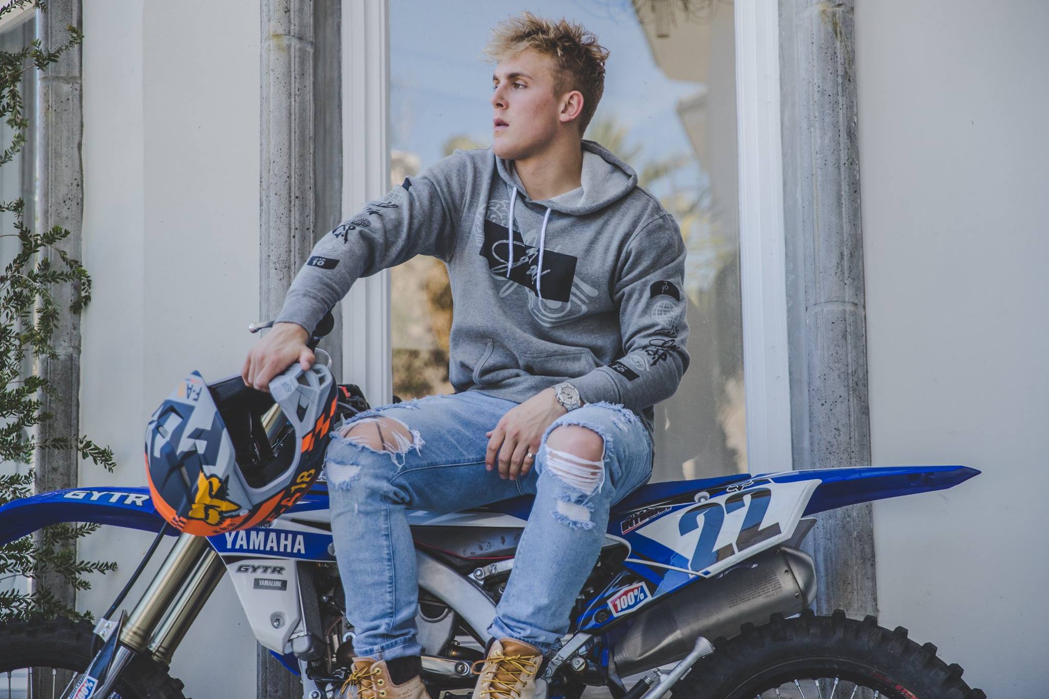 A young man in a hoodie sits on a blue motorcycle against a white wall, looking contemplative.