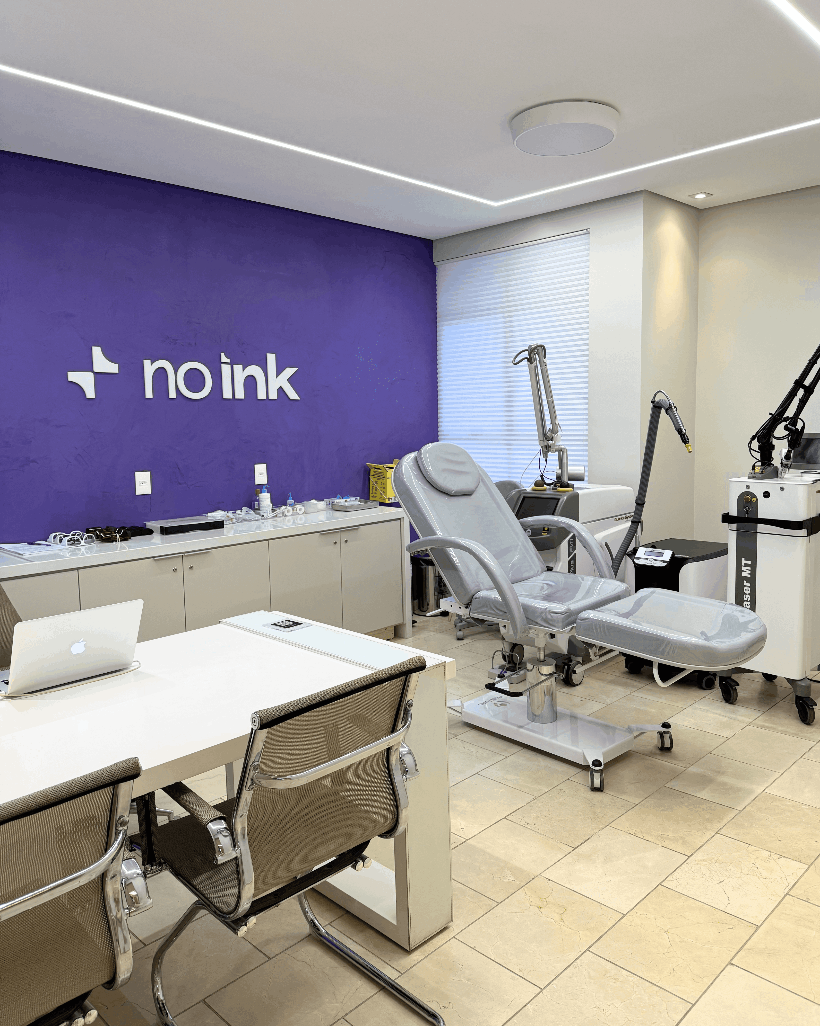 Consultório clínico com parede roxa exibindo a marca "NO INK", cadeira de procedimentos cinza, mesa com notebook e equipamentos médicos para remoção de tatuagem
