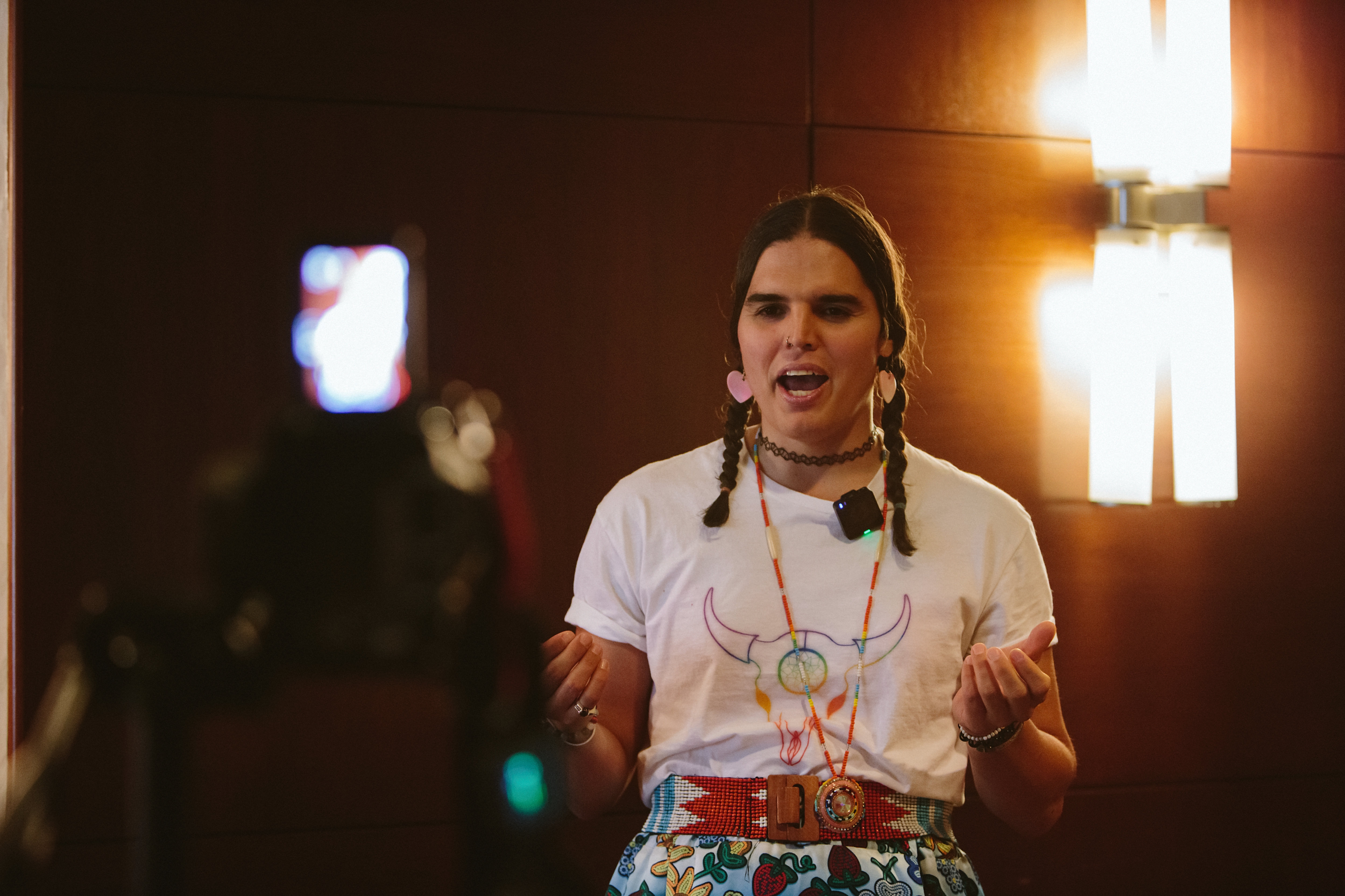 A person with long hair is speaking in front of a microphone, lit by warm wall lights, conveying a thoughtful message.