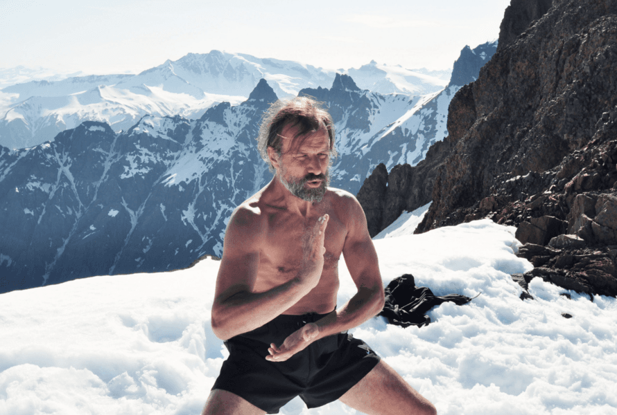 Wim Hof is meditating in a cross-legged position on a snowy, icy landscape, wearing minimal clothing, surrounded by large ice formations under an overcast sky, highlighting the contrast between the human form and the cold environment.