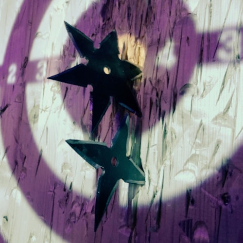 Duas estrelas de arremesso pretas embutidas em uma placa de alvo de madeira, iluminadas por luzes roxas e brancas.