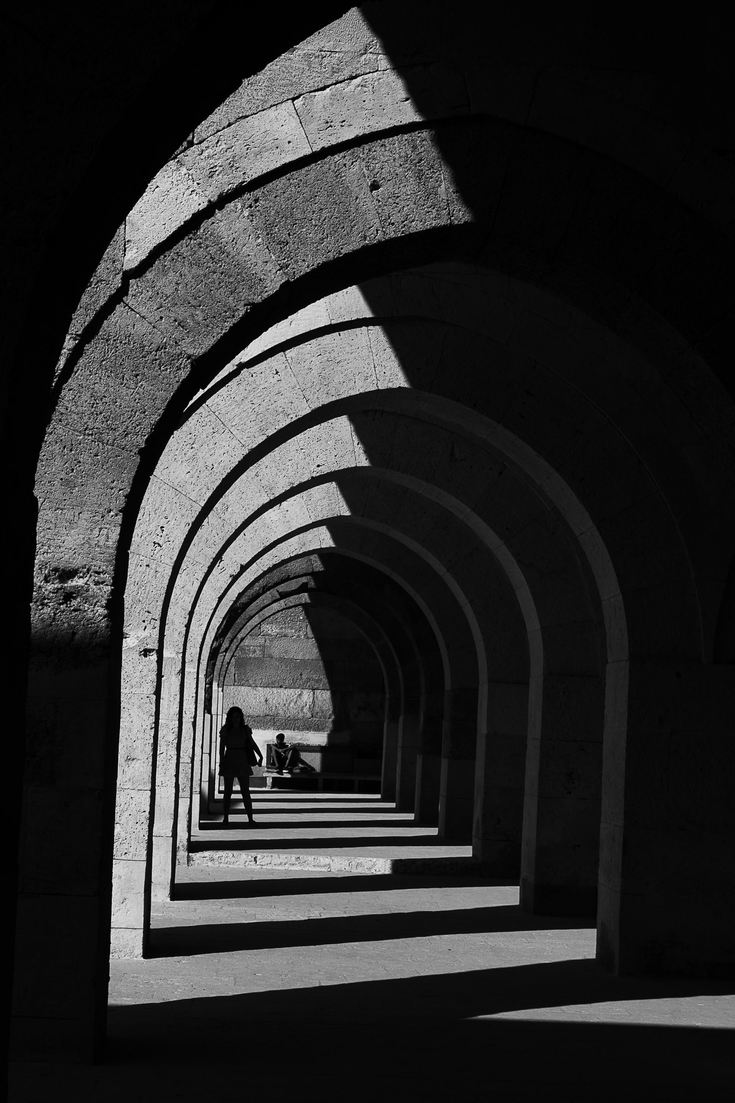 Stone archway corridor with strong light and shadow contrast, two figures framed in the distance.