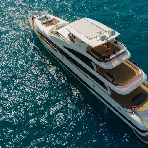 從高空俯瞰一艘大型白色遊艇在清澈藍色水面上航行，船上有幾個人。