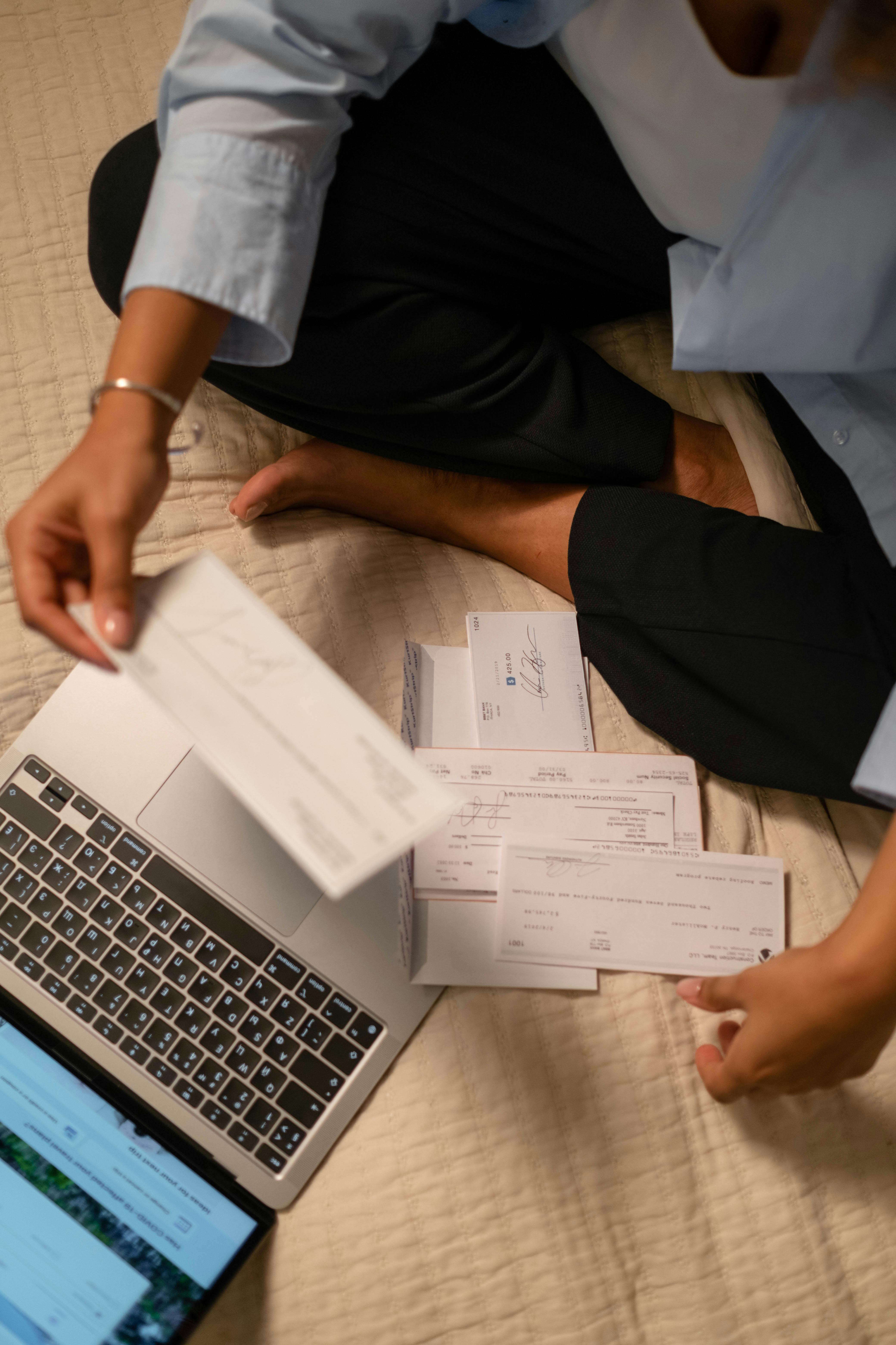 A person sits cross-legged while reviewing paper documents and working on a laptop, representing a hospital billing or accounts contact reaching out with questions about their anesthesia billing account