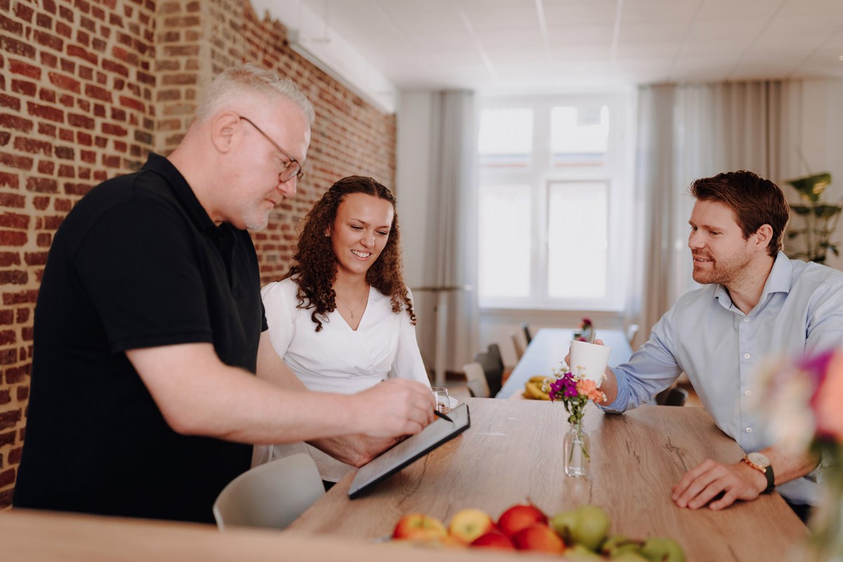 Weibliche Geschäftsführerin und zwei Personen sitzen gemeinsam an einer Bank und besprechen Unterlagen in einem hellen Büro von SLB.