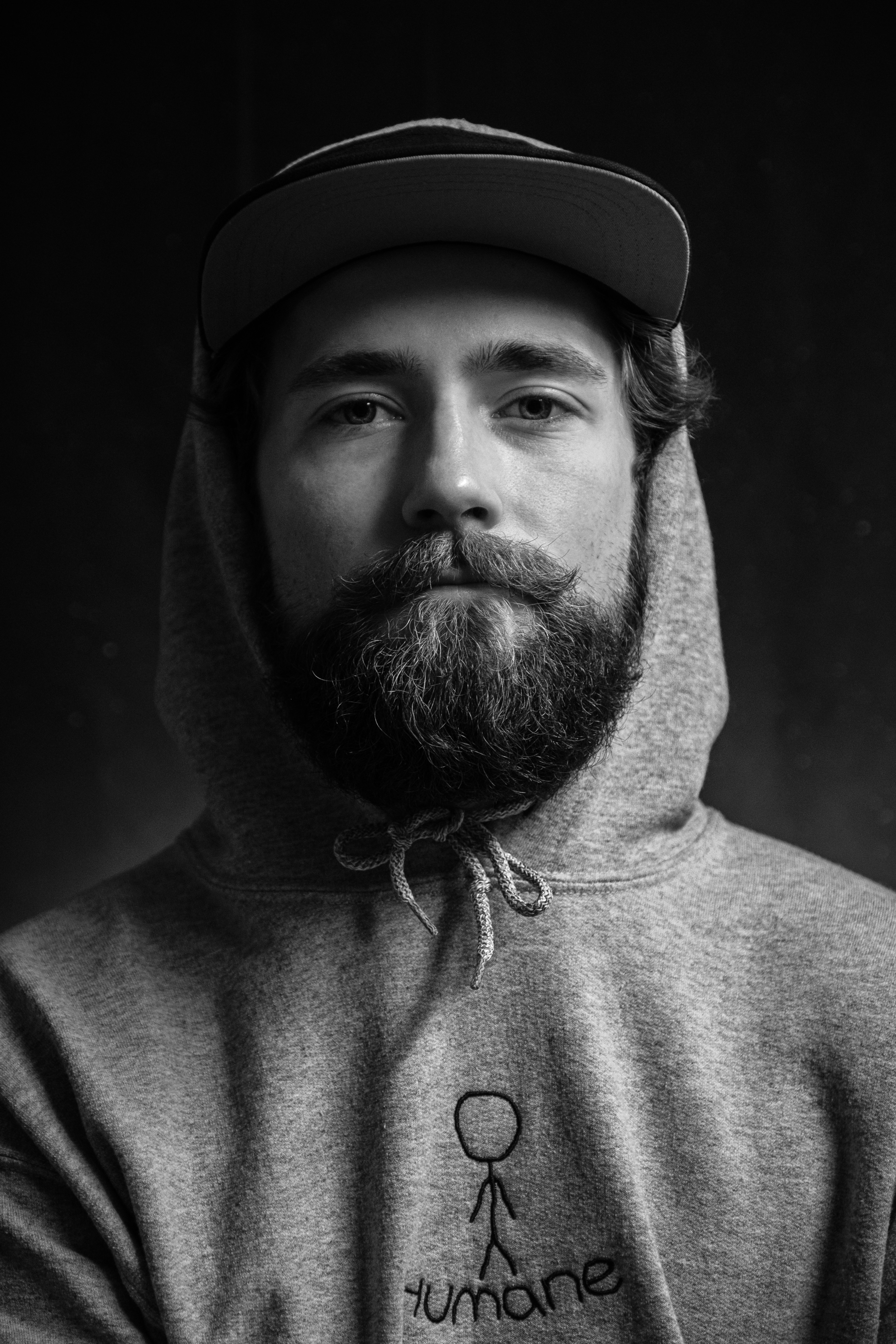 Black-and-white portrait of a bearded man wearing a hoodie and a cap, looking directly at the camera against a dark background.