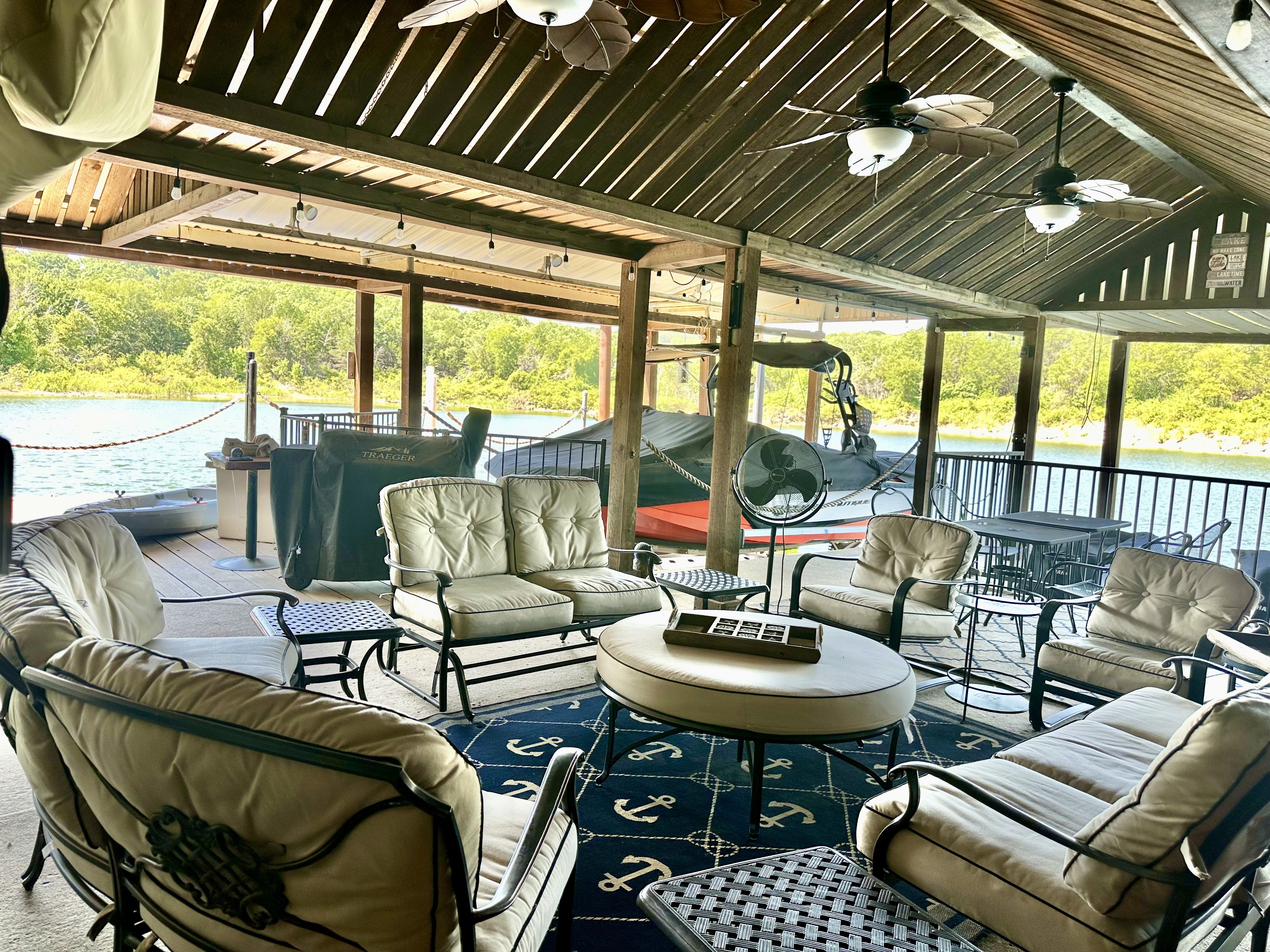 Outdoor patio with cushioned seating and circular tables under a wooden pergola, overlooking a serene lake with lush greenery.