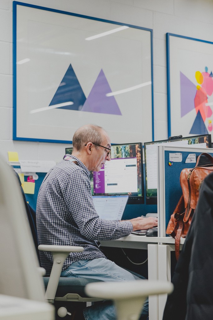 A person sits at a desk in an office space, working on a laptop with two computer monitors. The desk has a backpack hanging from the side, and the wall behind displays framed geometric artwork.