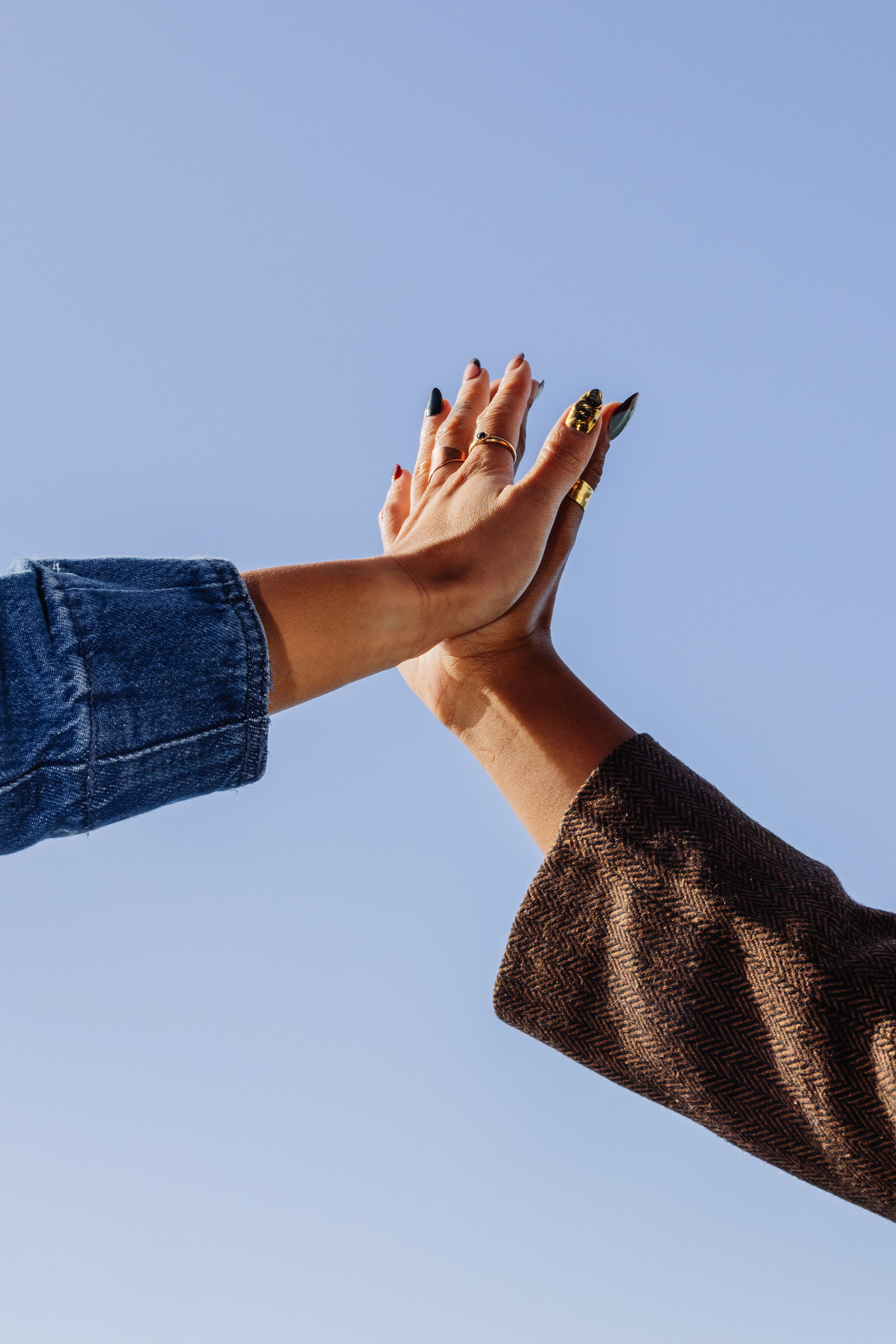 Two hands with rings and painted nails pressed together in a high-five gesture against a blue sky background.