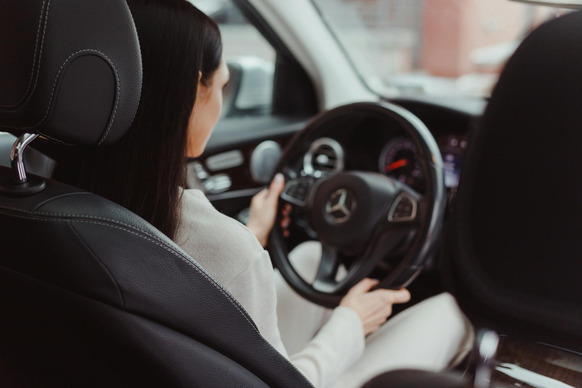  An over-the-shoulder shot from the back seat showing a woman with long, dark hair driving a modern Mercedes-Benz (indicated by the steering wheel logo). She is wearing a light-colored top and gripping the steering wheel. The interior features black leather seats and the view through the front window is blurred, suggesting the car is in motion.