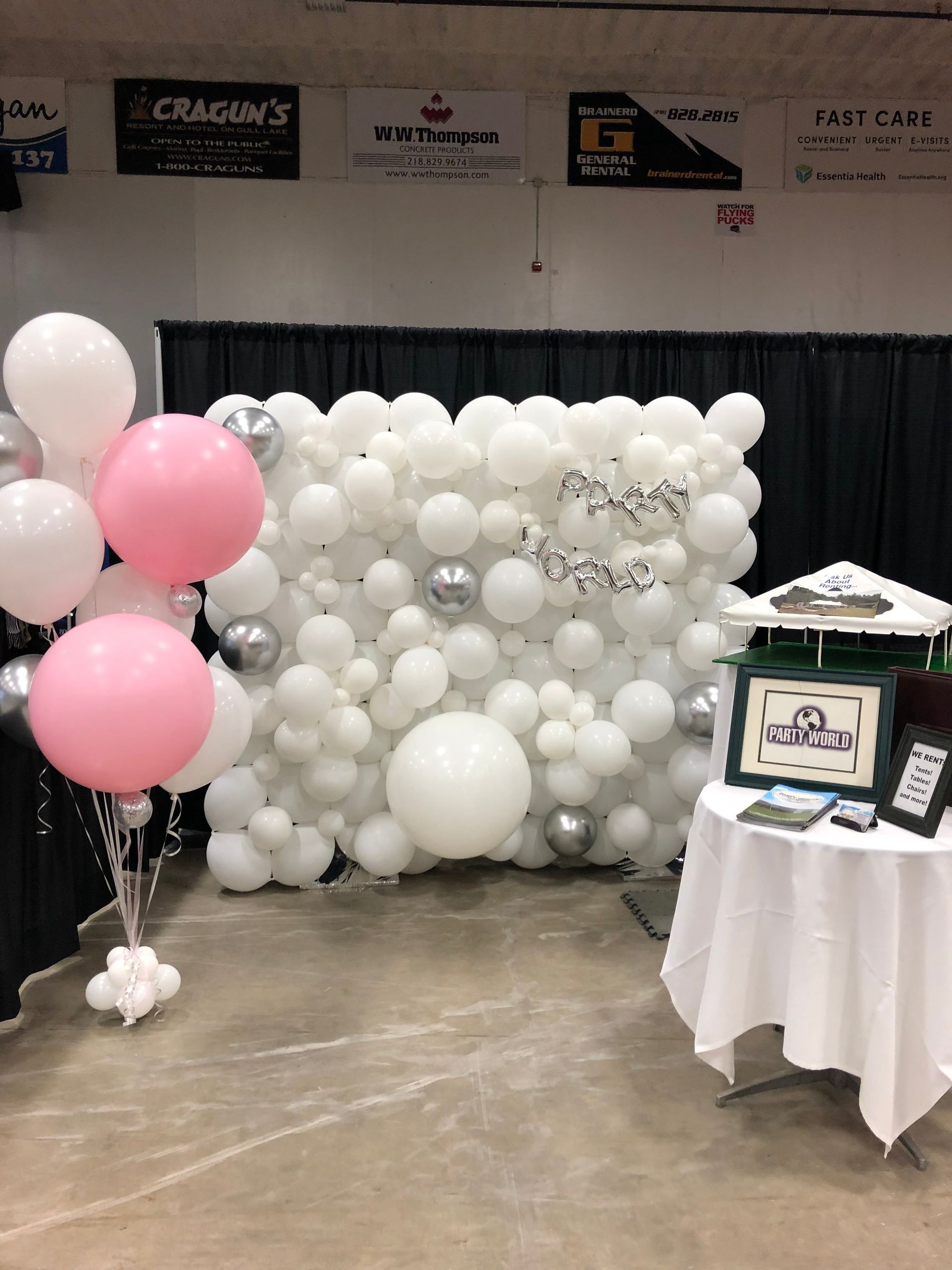 A photo of a festive event setup featuring a wall of white and silver balloons, with a decorative arrangement of pink and white balloons on the side, alongside a table with informational brochures and a small poster for Pet World, set against a backdrop of black curtains with overhead advertisements.