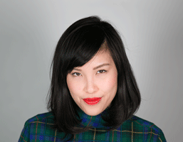The image presents a portrait of a young woman with shoulder-length dark hair, styled with a side part and bangs that fall across her forehead. She is wearing a dark turtleneck top with a plaid pattern in shades of green, blue, and possibly yellow or gold. The background is a plain light gray or off-white color. Her gaze is directed straight at the camera, and she has red lipstick on her lips.
