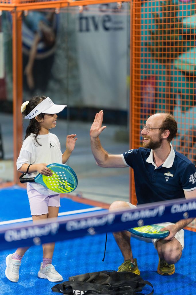 A young girl wearing a white cap and a t-shirt smiles and high-fives a man wearing a navy blue polo shirt, both holding paddles on a blue indoor court.
