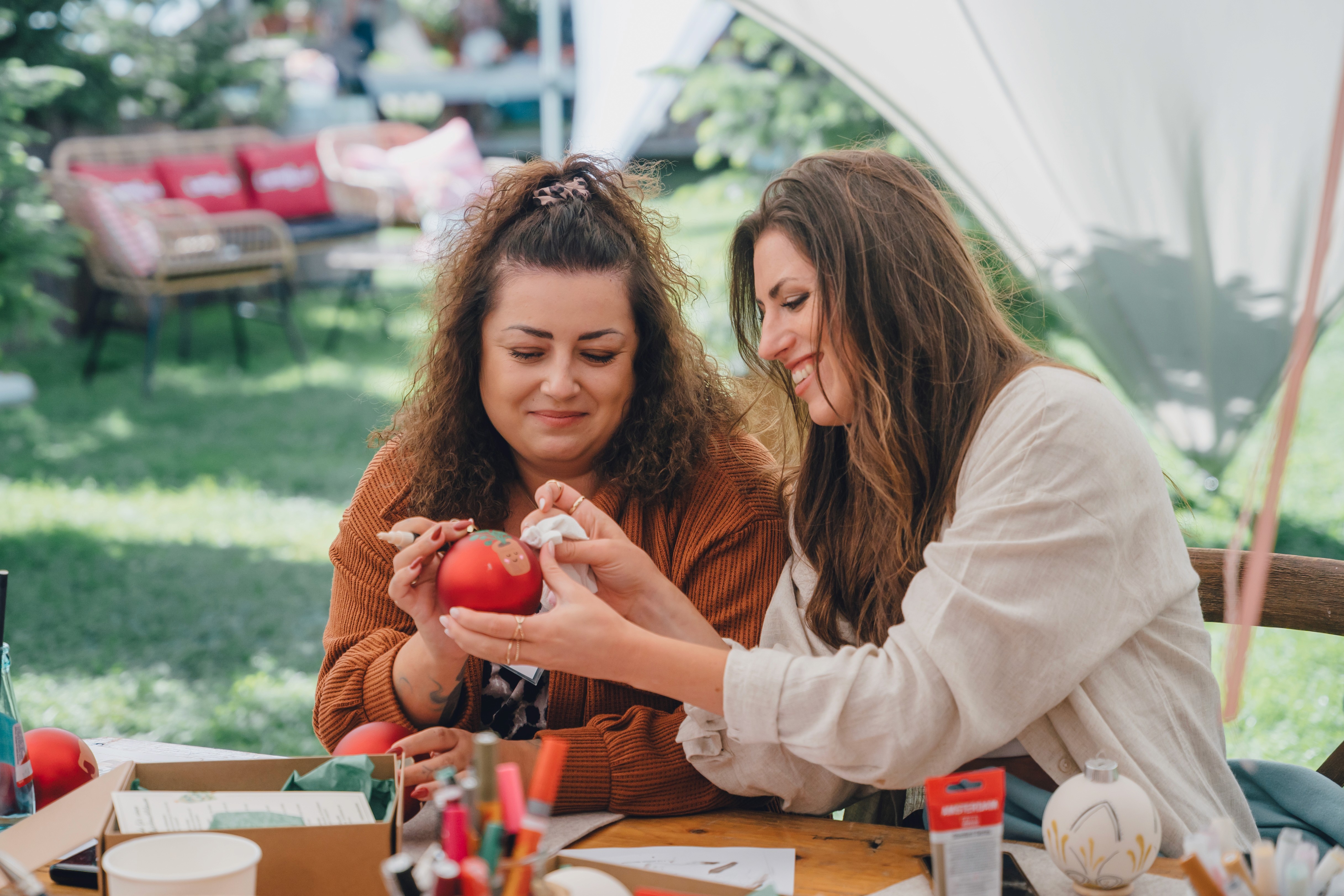 Zwei Frauen gestalten Weihnachtsbaumdekoration bei einem DIY-Workshop an einem Holztisch im geschmückten Zelt. 