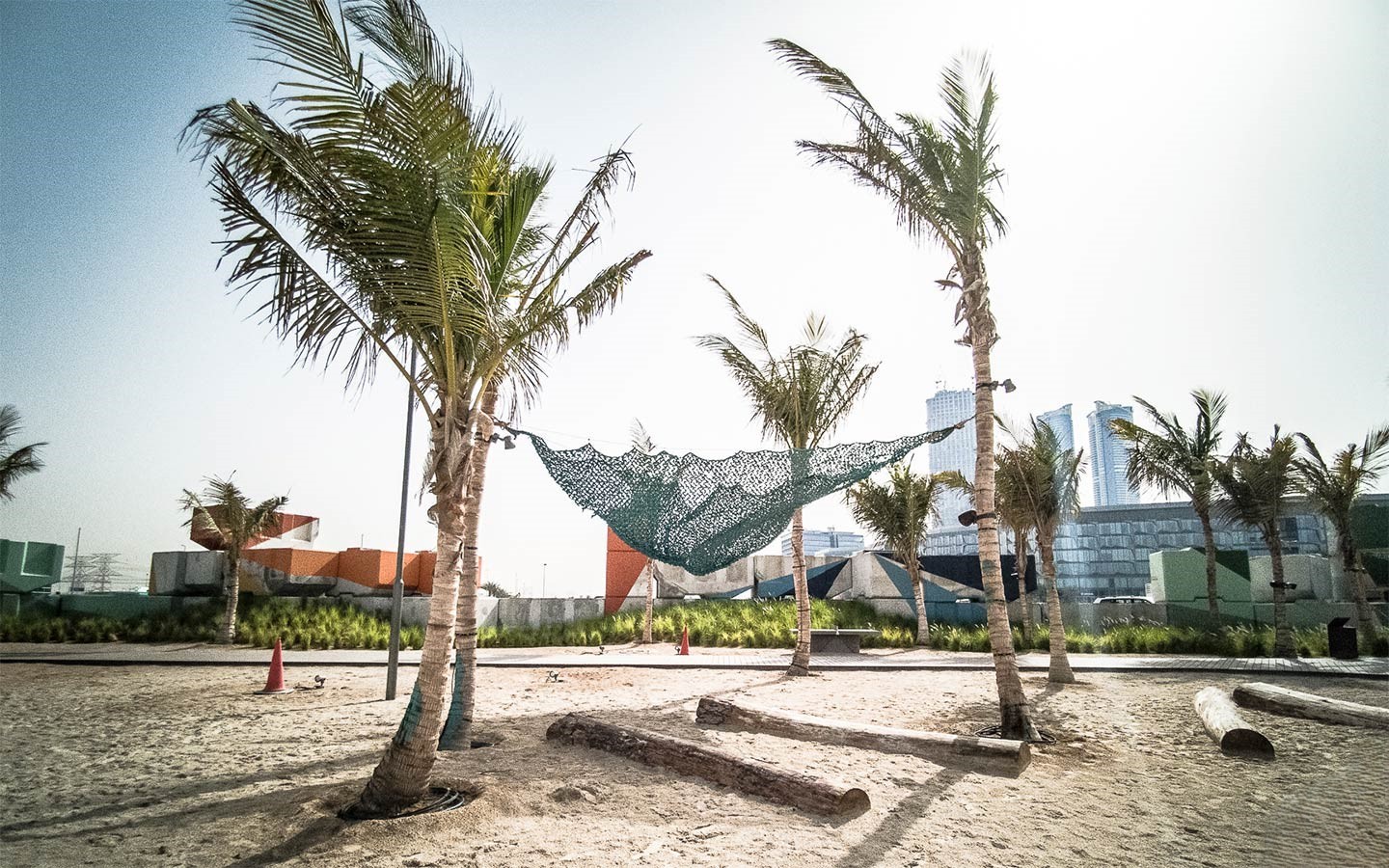 A small, sandy recreational area with palm trees, a hammock made of camouflage netting, and modern buildings of Dubai Design District in the distance.