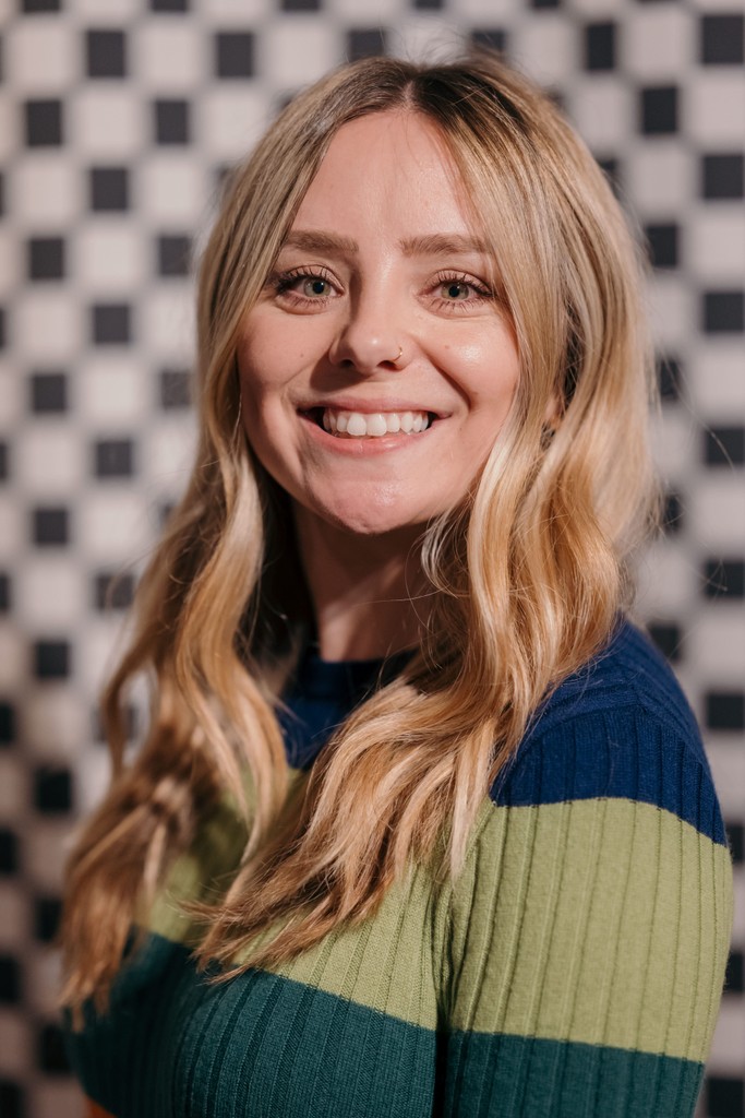 A person with long wavy blonde hair is shown from the shoulders up, wearing a striped blue and green sweater. The background features a black‑and‑white checkerboard pattern.