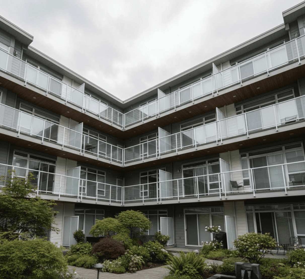 Townhouse strata courtyard looking up at multiple balconies with identical frameless glass railings, just clean sheets of glass on every unit, seamless transparent barriers, landscaped common area below, overcast Vancouver sky