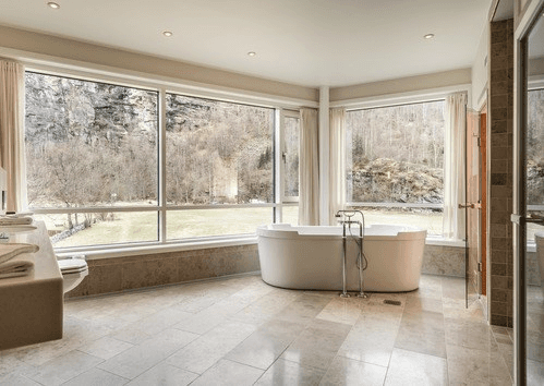 A luxurious bathroom features a freestanding white bathtub near large, bright windows offering a scenic view of a rocky landscape and sparse trees, complemented by beige marble flooring and soft, neutral-toned decor.