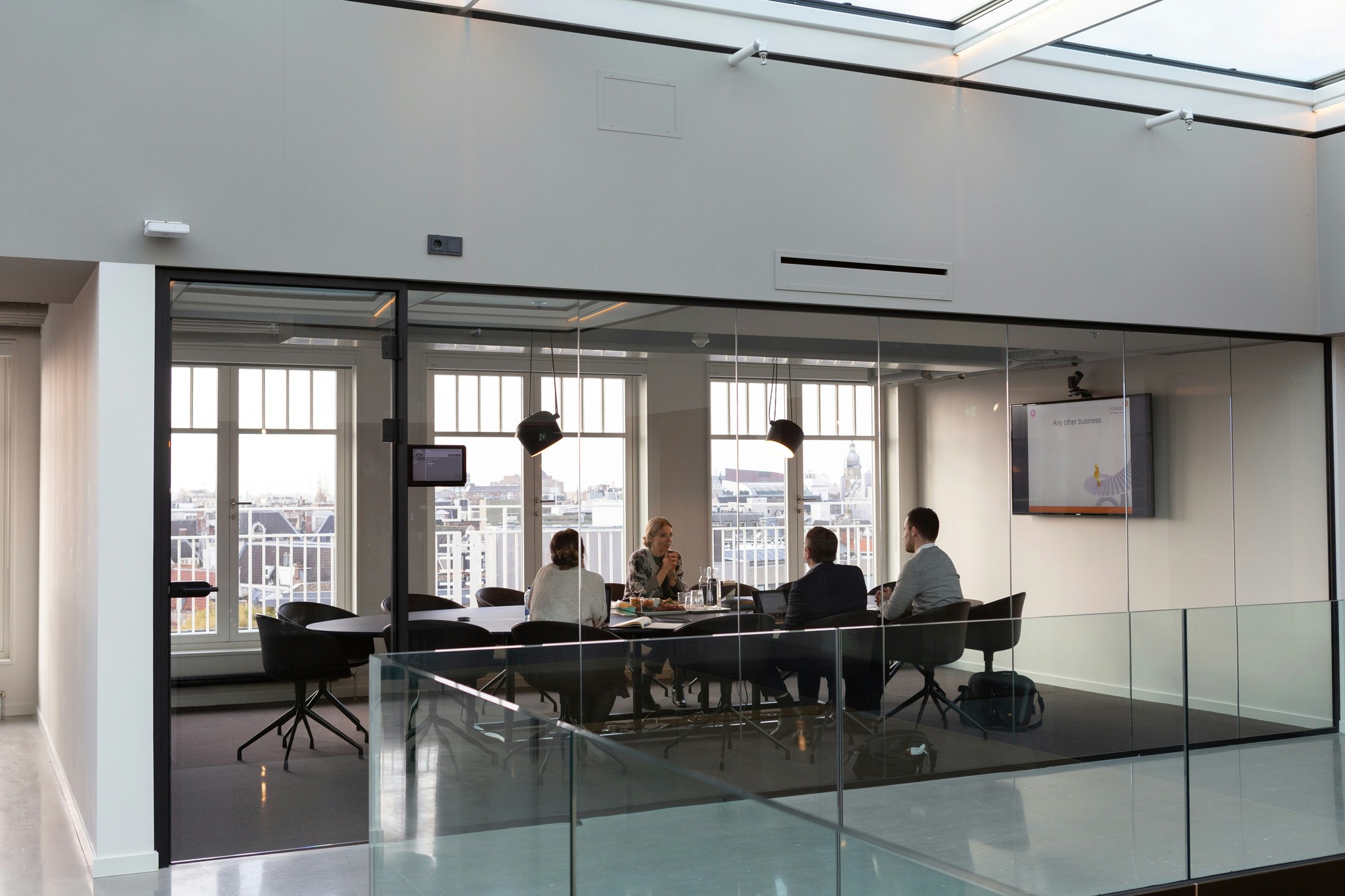 A modern office meeting room with large windows overlooking an urban landscape and people gathered around a conference table.