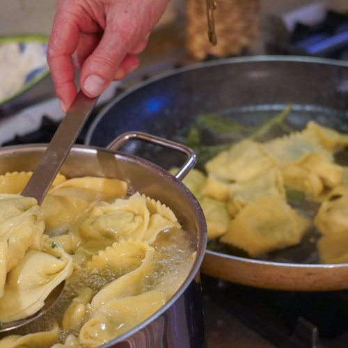 A person uses a ladle to scoop steaming ravioli from a pot of boiling water on a stovetop. Another pan is seen in the background.