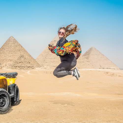A woman jumps joyfully in front of the Pyramids of Giza under a clear sky, with an ATV parked to the side.