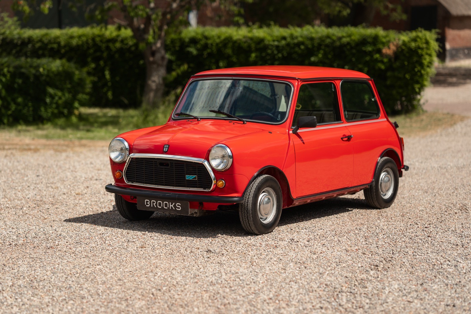 A bright red compact car parked on a gravel road, surrounded by lush green hedges and trees.