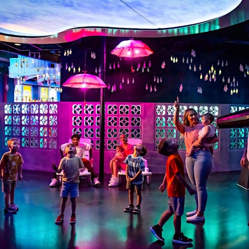 Children and adults look up in a brightly lit room with colorful lights, hanging pink umbrellas, and decorative wall panels.