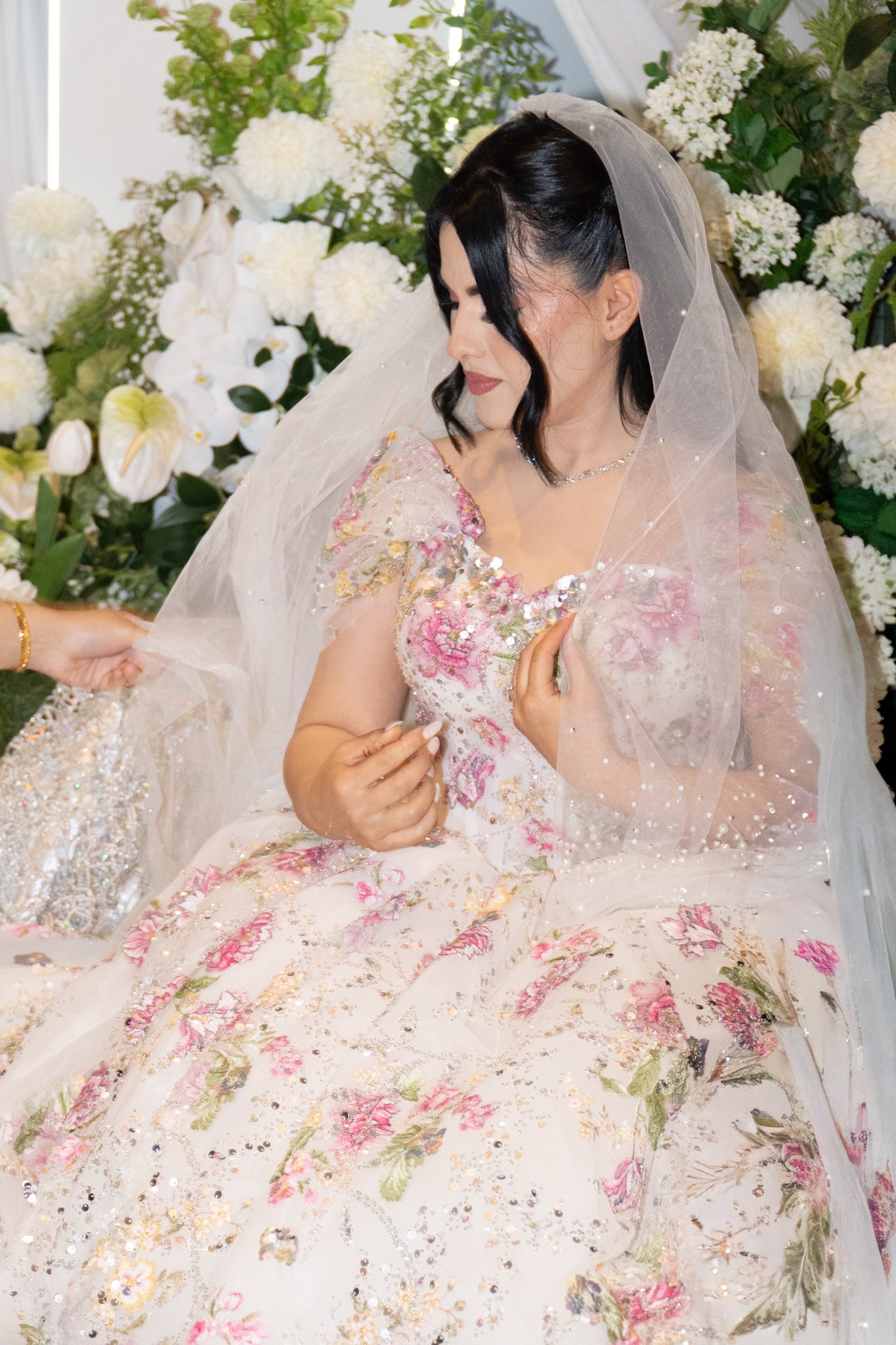 Bride in a floral wedding gown and veil seated beside an arrangement of white flowers.