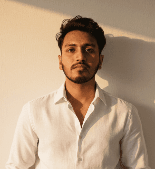 A man wearing a white shirt poses in front of a simple wall, with a neutral expression on his face.