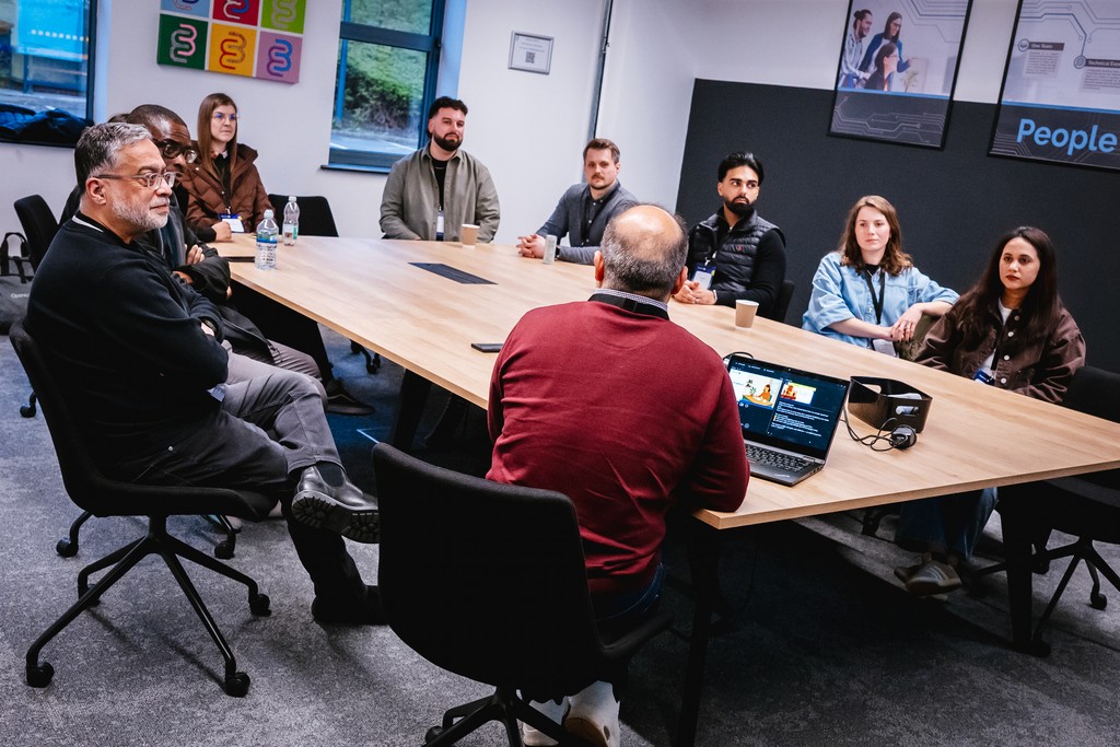 A group of people sit around a long conference table in a modern office, listening to a seated speaker with a laptop open in front of them. Cups, water bottles, and a headset are on the table, and posters and windows are visible on the walls behind.