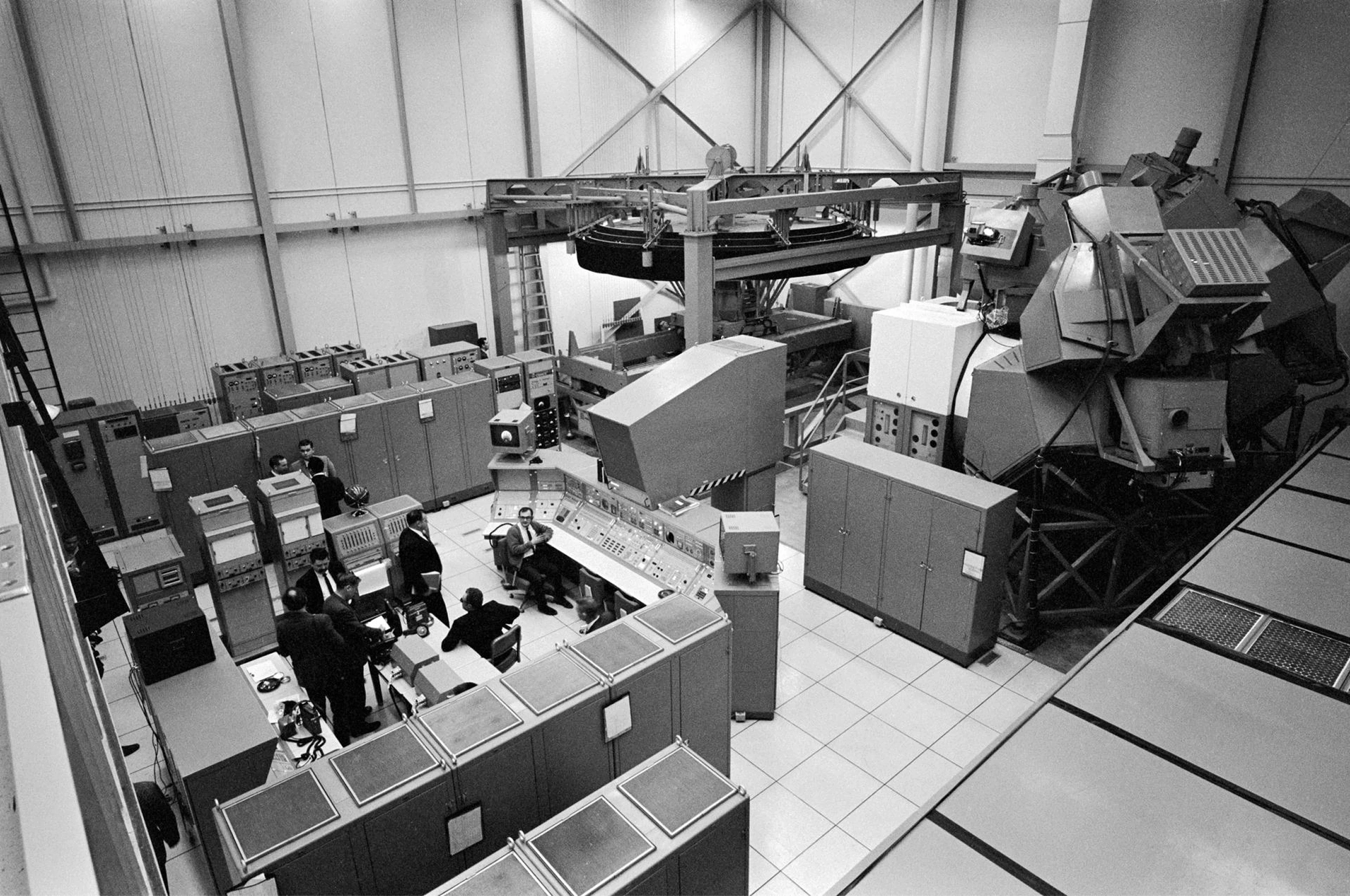 Overhead black and white photograph of a NASA simulator facility, showing a large hardware rig surrounded by banks of control consoles. Photo: NASA