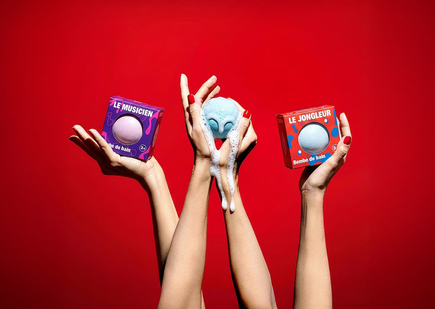 Bombes de bain artisanales colorées sur fond rouge, présentées à la main : une bombe de bain moussante bleue et deux bombes de bain emballées nommées Le Musicien et Le Jongleur, concept créatif et ludique inspiré de l’univers du cirque.